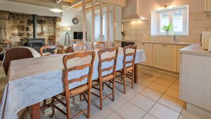 A dining area with a marble table and wooden chairs, a stone fireplace, and a kitchen with white cabinets and a window. - Gite Le Moutat-La Cave with Private Garden and Wi-Fi (Allemans)