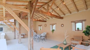 Un interno rustico con travi a vista in legno, una zona pranzo con sedie bianche e un angolo salotto con arredi in legno.