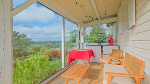 Un patio techado con una mesa y sillas, un mantel rojo, y vistas a zonas verdes y colinas.