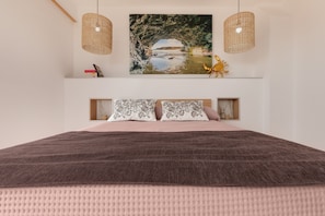 A bedroom with a large bed, two bedside lamps, a picture of a beach scene, and a small shelf with books and a decorative item.