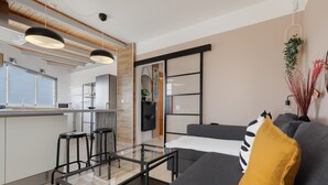 Een moderne woonkamer met een glazen salontafel, een grijze bank met een mosterdkleurig kussen, en een keuken in de achtergrond.