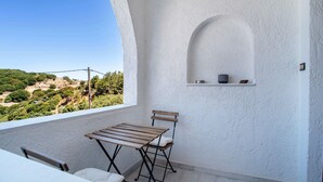 A balcony with a table and chair, a white wall with an arched niche, and a view of a green landscape.