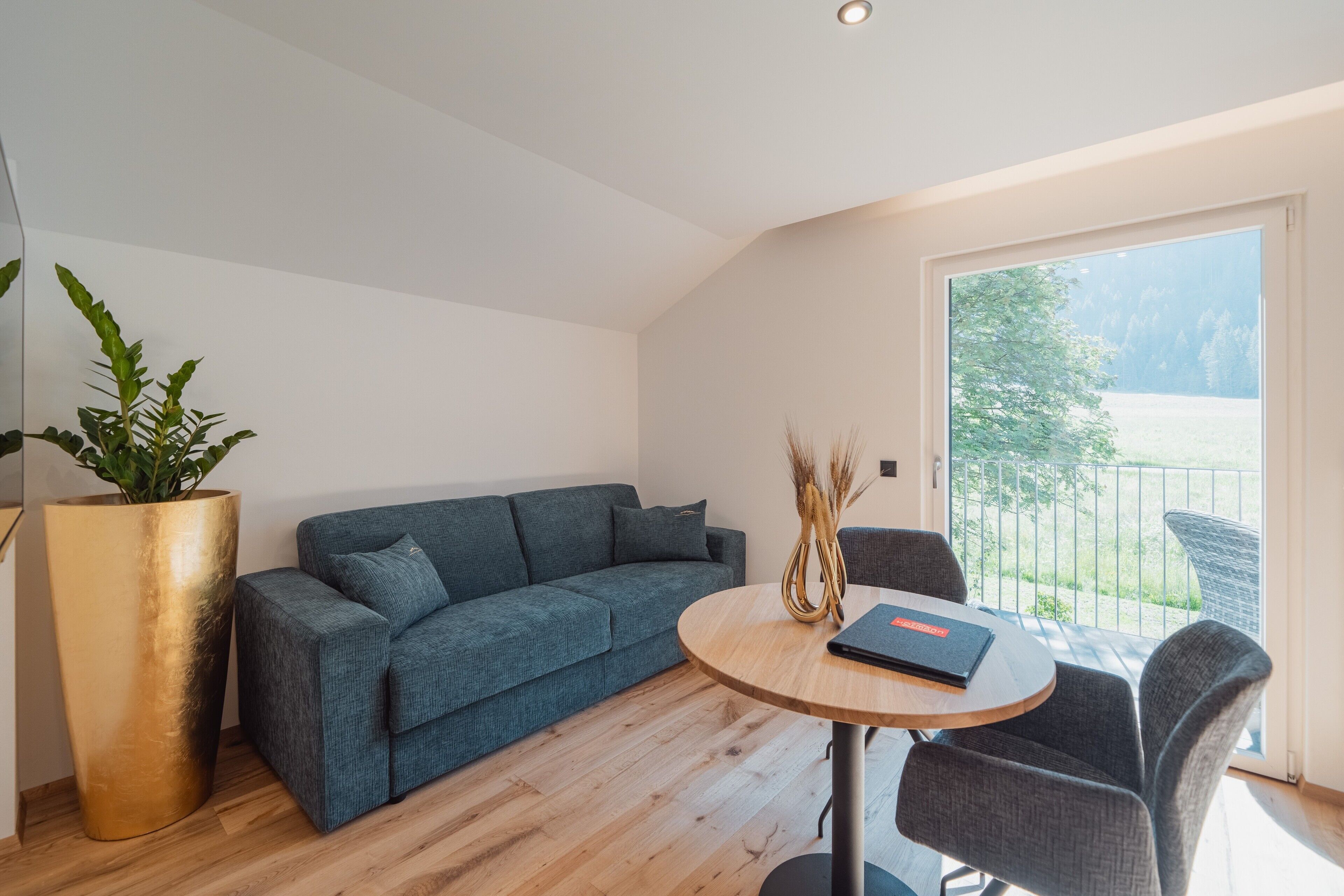 Ein modernes Wohnzimmer mit einem blauen Sofa, einem runden Holztisch und einem großen Fenster mit Blick ins Grüne.
