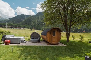 A wooden cabin with a small porch, a sofa, and a barbecue grill in a grassy area with mountains in the background. - Apartment 'Alpenrose' with Mountain View, Shared Terrace and Wi-Fi (St. Martin in Gsies)
