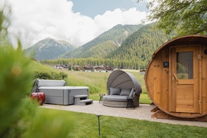A wooden cabin with a covered seating area, a hot tub, and a view of mountains.