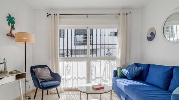 A modern living room with a blue sofa, a round coffee table, a desk with a lamp, and a view through a large window. - Apartment 'Blue Beach Home' with Terrace (Conil de la Frontera)