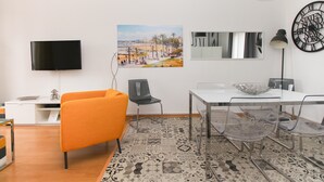 Una habitación moderna con televisión de pantalla plana, un cuadro con escena de playa, un reloj y un comedor con sillas transparentes y una mesa blanca.