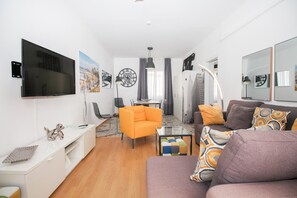 Uma sala de estar moderna com televisão de tela plana, uma mesa de centro de vidro, uma poltrona laranja, um sofá com almofadas estampadas e um espaço de jantar com lustre.