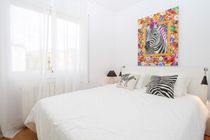 Une chambre avec un grand lit, du linge de lit blanc, une œuvre d’art sur le thème des zèbres accrochée au mur et une fenêtre ornée de rideaux légers.