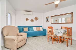Ein Wohnzimmer mit einer Couch, einem Sessel, einem Esstisch mit Stühlen, einem Deckenventilator, Wandklimaanlage und einem Spiegel.