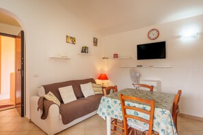 Een woonkamer met een bank, een eettafel met stoelen, een aan de muur bevestigde televisie en een klok.