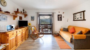 A living room with a sofa, a rocking chair, a TV, wooden cabinets, and a window with a view. - Apartment 'Sylvie' with Mountain View, Shared Terrace and Wi-Fi (Gressan)