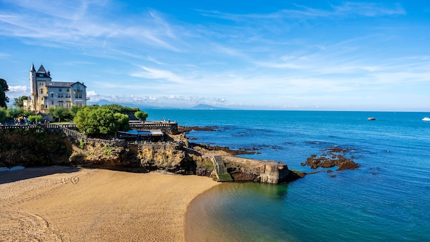 A coastal hotel with a beach, clear blue waters, and a mountainous backdrop.