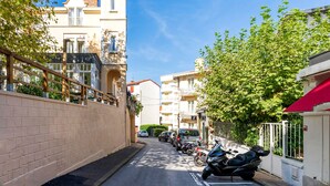 A narrow street lined with parked cars and motorcycles, buildings with balconies, and a clear blue sky.