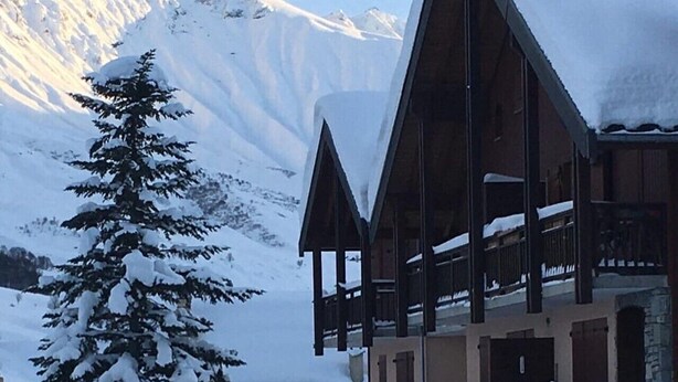 Un chalet dont l’extérieur est recouvert de neige, avec une structure en bois imposante, et qui se dresse sur fond de montagnes.