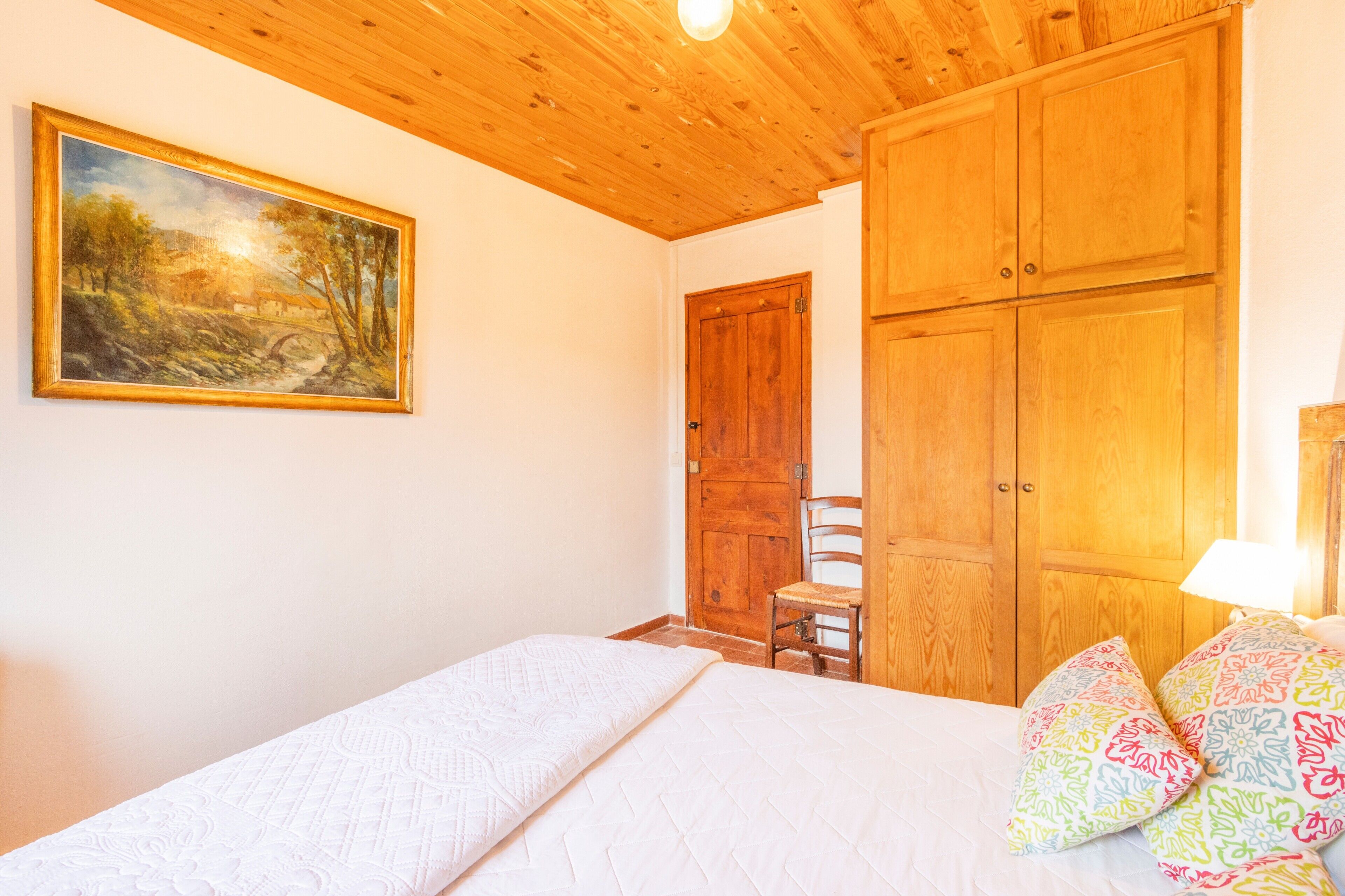 A bedroom with a bed, a wooden wardrobe, a chair, and a painting on the wall.