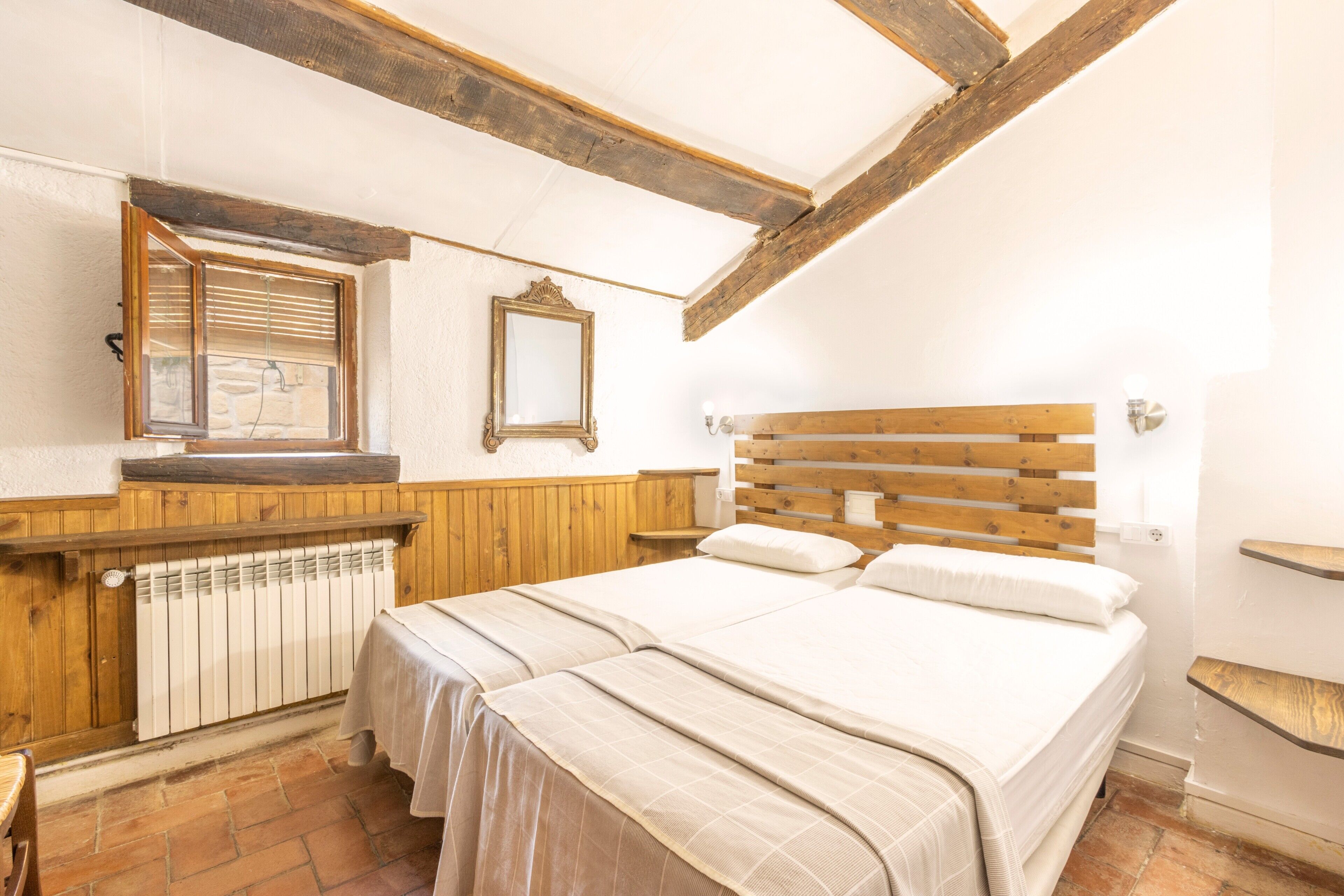 Ein Schlafzimmer mit einem hölzernen Kopfteil, einem Bett mit zwei Kissen, einer Holzbank, einem Heizkörper und einem Spiegel an der Wand.