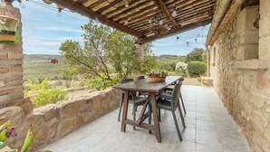 A rustic outdoor patio with a wooden table and chairs, a stone wall, and a view of the countryside.