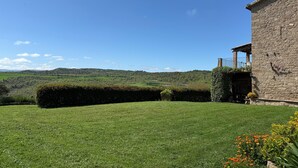 A stone building with a garden, a clear blue sky, and a hilly landscape in the background.