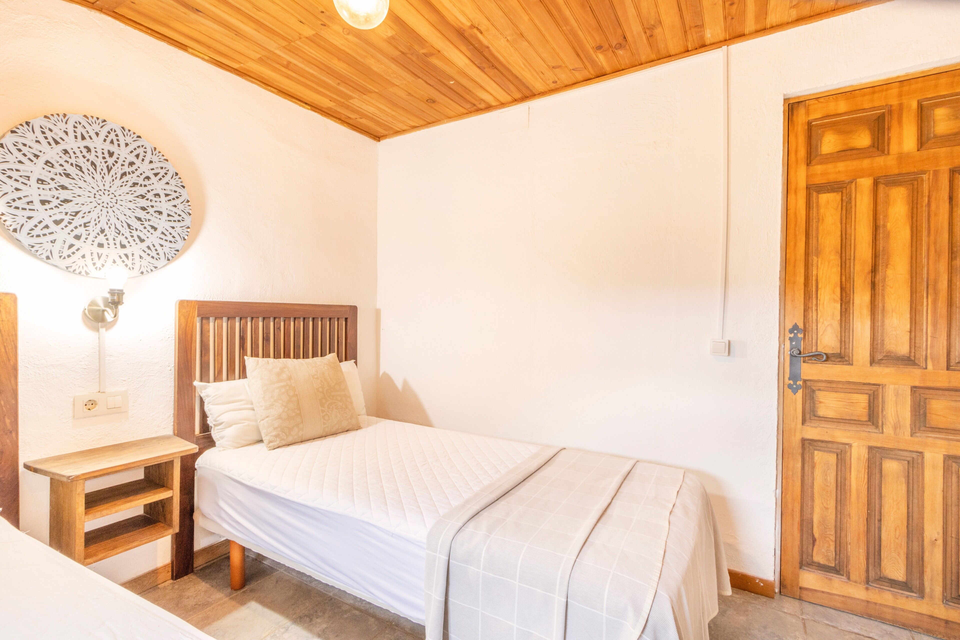 A bedroom with a wooden headboard, a bedside table, a bed with a quilted blanket, a decorative wall piece, and a wooden door.
