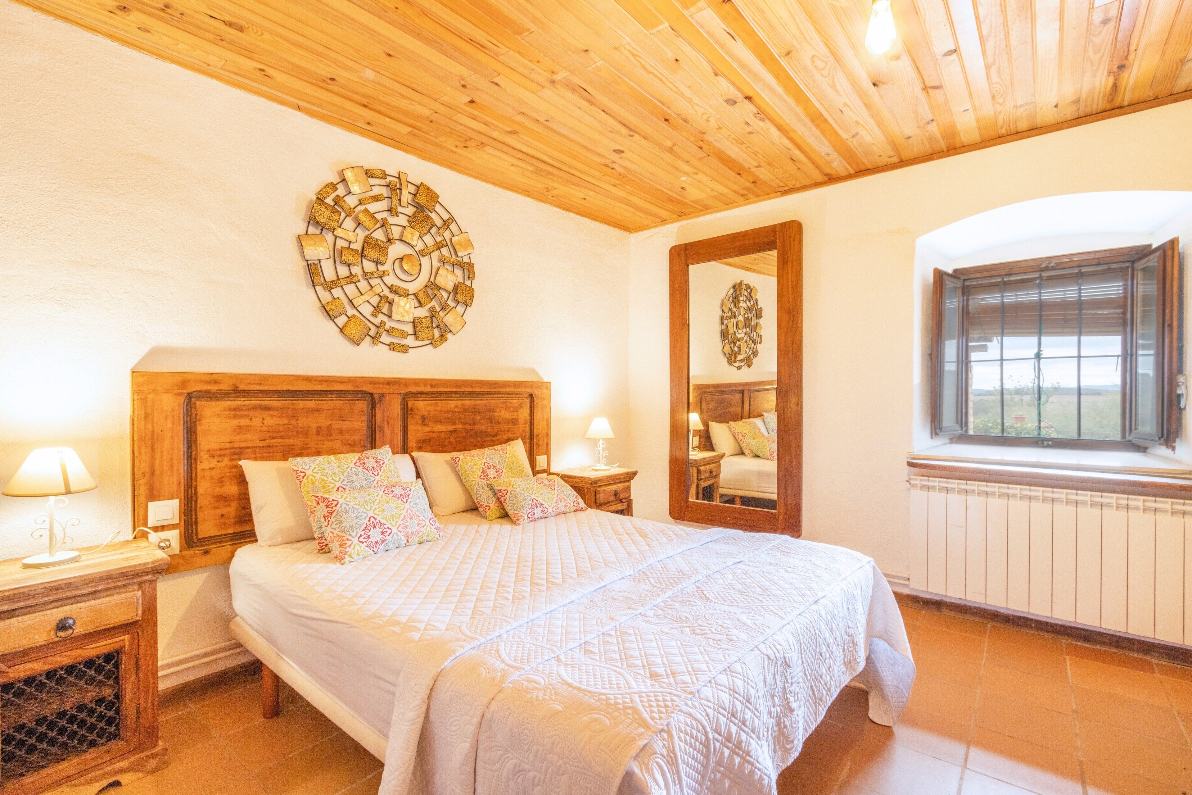 A bedroom with a wooden ceiling, a large bed with patterned pillows, a wall clock, and a window with a view.
