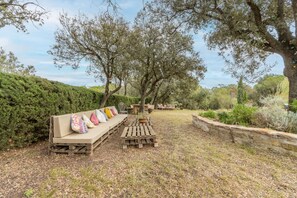 An outdoor seating area with a sofa and a wooden table, surrounded by trees and a stone wall.