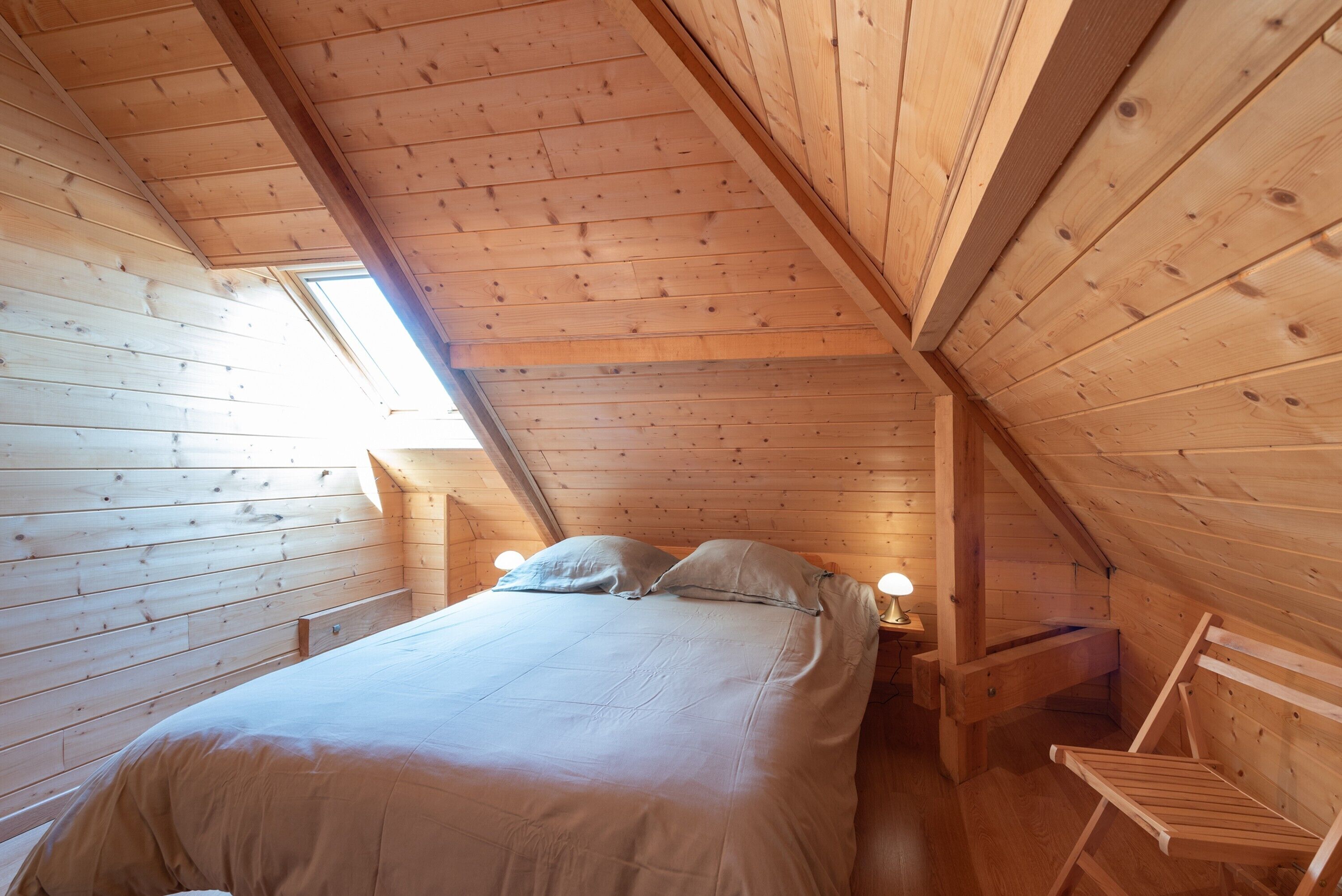 A wooden cabin bedroom with a bed, two bedside lamps, a chair, and a skylight.