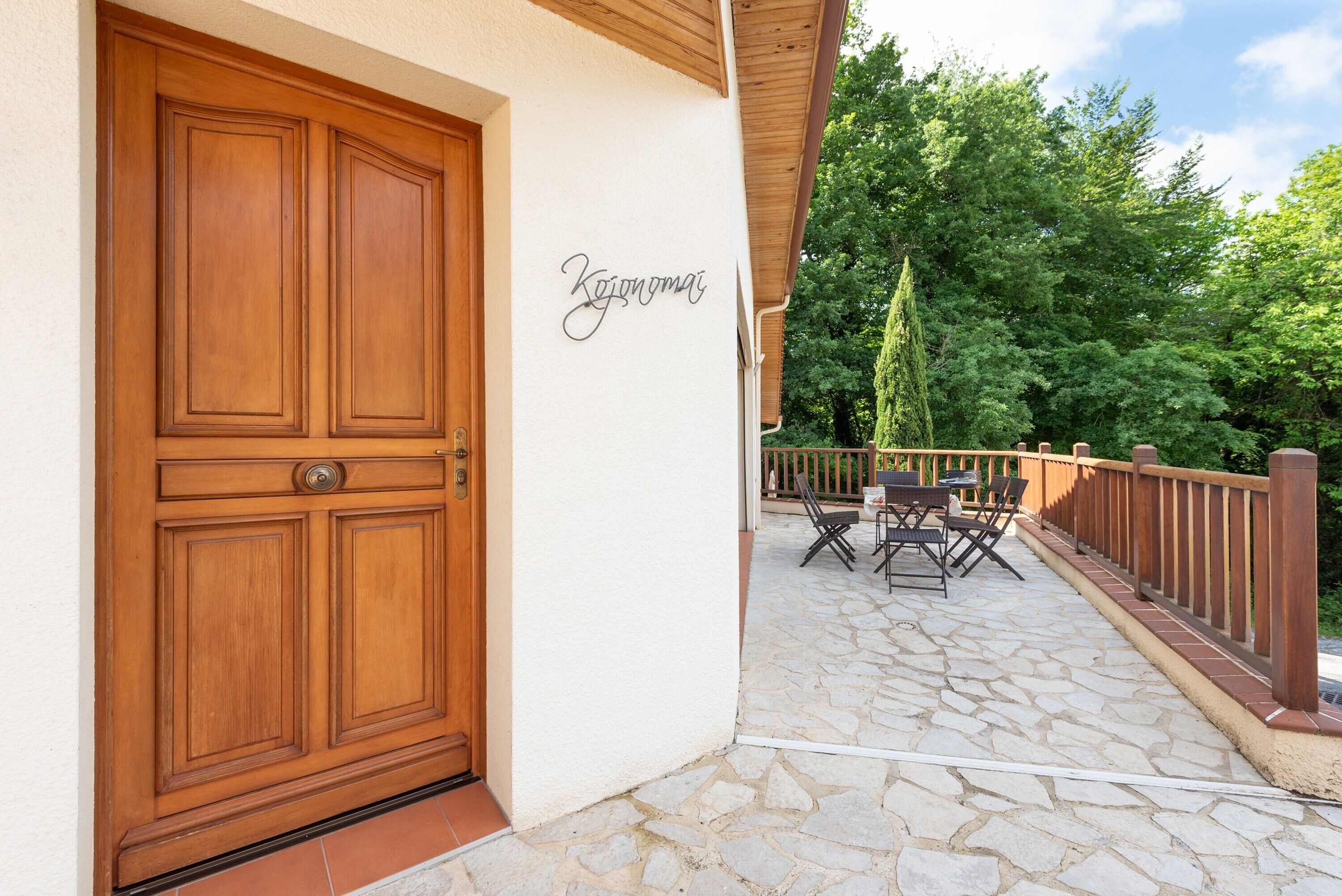 Une porte en bois avec une plaque nominative, une terrasse en pierre, du mobilier d’extérieur et une rambarde en bois.
