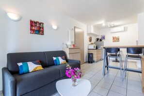 Een moderne woonkamer met een bank, een bijzettafeltje met bloemen, een eethoek met stoelen, en een keuken op de achtergrond.