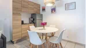A modern kitchen with wooden cabinets, a white dining table with chairs, a hanging light fixture, and wall-mounted pictures. - 'Meu Descan' Apartment with Shared Pool, Private Terrace, and Air Conditioning (Cerbère)