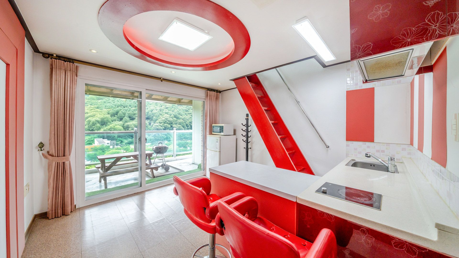 A modern kitchen with a red and white color scheme, a dining area with red chairs, and a view of greenery through the sliding glass door.