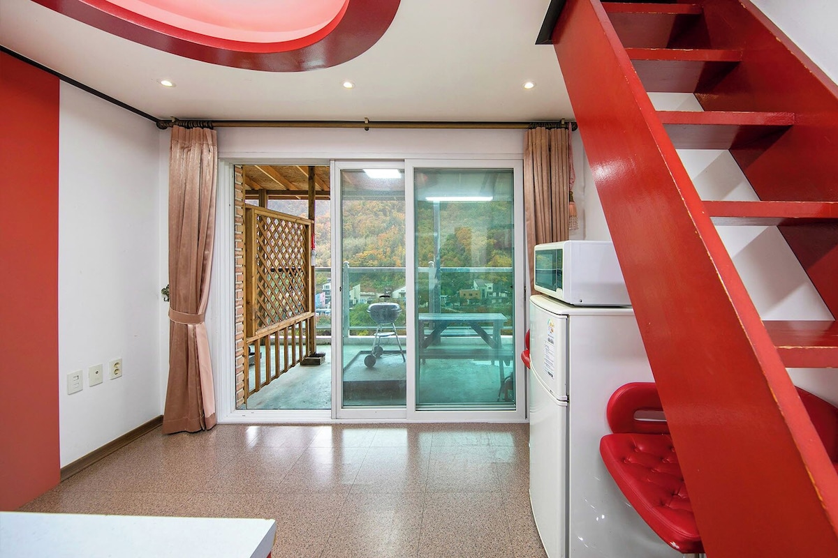 Une pièce avec un escalier rouge, une porte-fenêtre coulissante donnant sur un balcon et un petit coin cuisine.