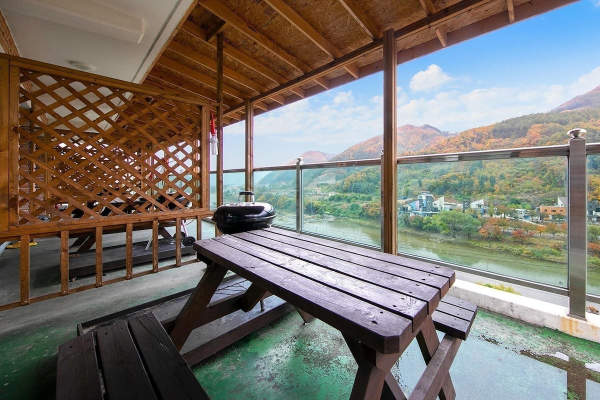 Eine Holzterrasse mit Picknicktisch und Grill, mit Blick auf einen Fluss und herbstliche Hügel.