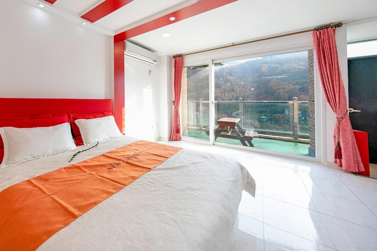 Een slaapkamer met een groot bed, een rode hoofdbord en uitzicht op een balkon met een tafel en stoelen.