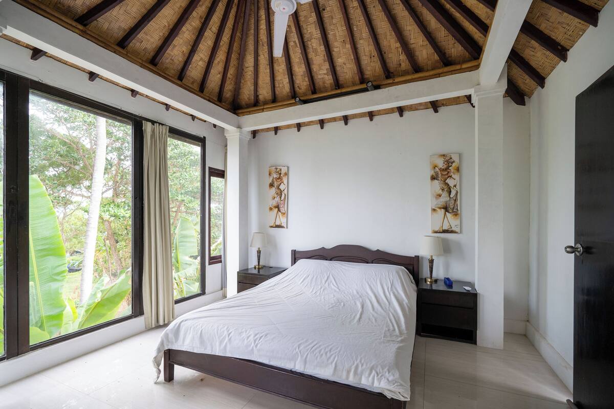 Ein Schlafzimmer mit einem großen Bett, einem Holz-Kopfteil, zwei Nachttischen mit Lampen und Blick auf Grün hinter großen Fenstern.