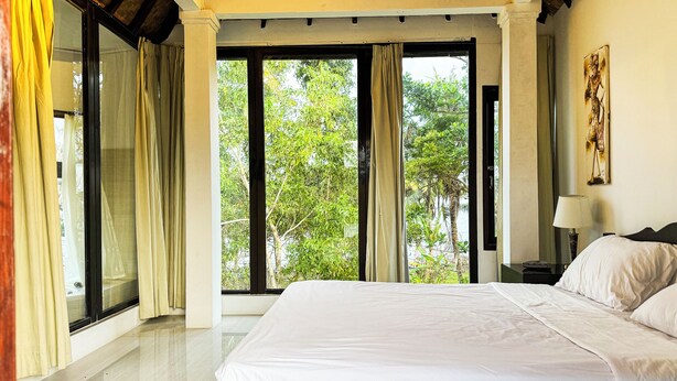 A hotel room with a large bed, a nightstand with a lamp, and a view of greenery through the windows. - Seva Hotel (Karangasem)