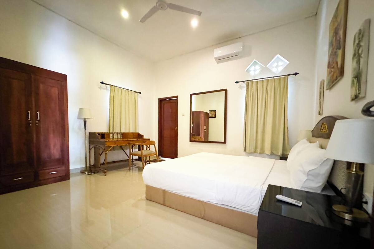 Ein Hotelzimmer mit einem großen Bett, einem Schreibtisch mit Stuhl, einem Kleiderschrank, einem Deckenventilator, einer Klimaanlage und einem Fenster mit Vorhängen.