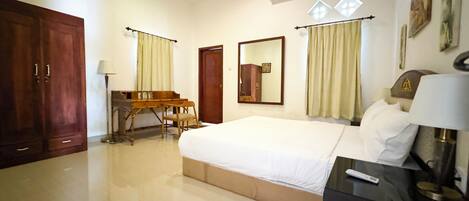 A hotel room with a large bed, a desk with a chair, a wardrobe, a ceiling fan, an air conditioning unit, and a window with curtains.