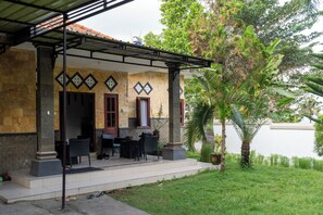 A covered outdoor seating area with chairs and a table, a door, and a small garden with a palm tree. - Seva Hotel (Karangasem)