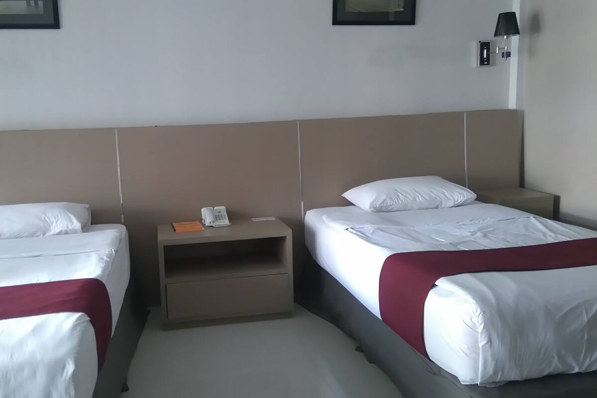 A hotel room with two single beds, each with white bedding and a red blanket. There is a small bedside table with a phone and a notepad. The room has a neutral color scheme with beige walls and a white ceiling.