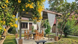A covered outdoor seating area with a table and chairs, surrounded by greenery and a tree with yellow flowers.