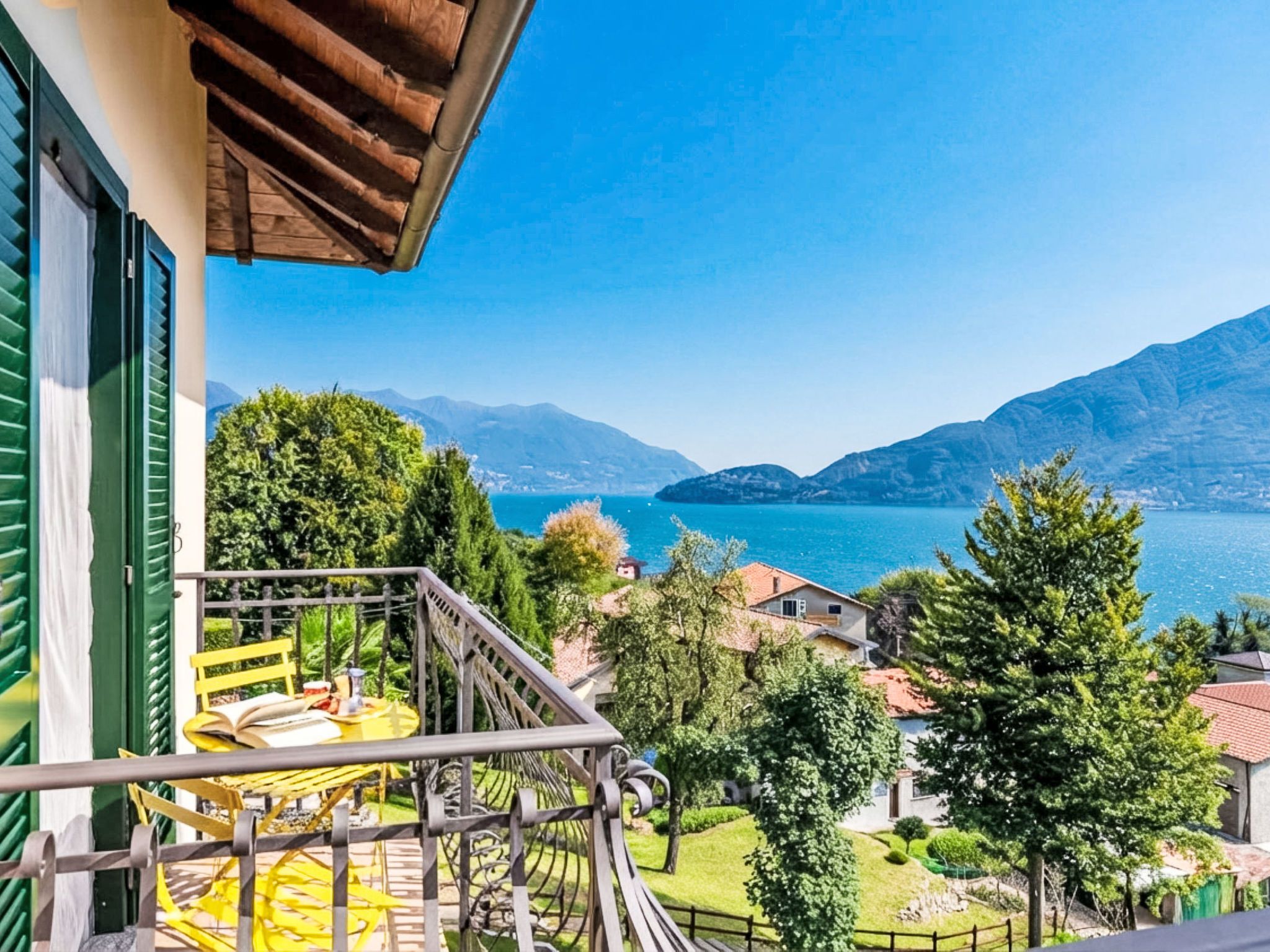 A balcony with a view of a lake and mountains, a dining area with outdoor furniture, and a garden with trees.
