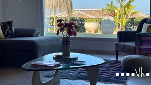 Een woonkamer met een glazen salontafel, een bank en een stoel. Er is uitzicht op een tuin met een grote pot en een hangplant.