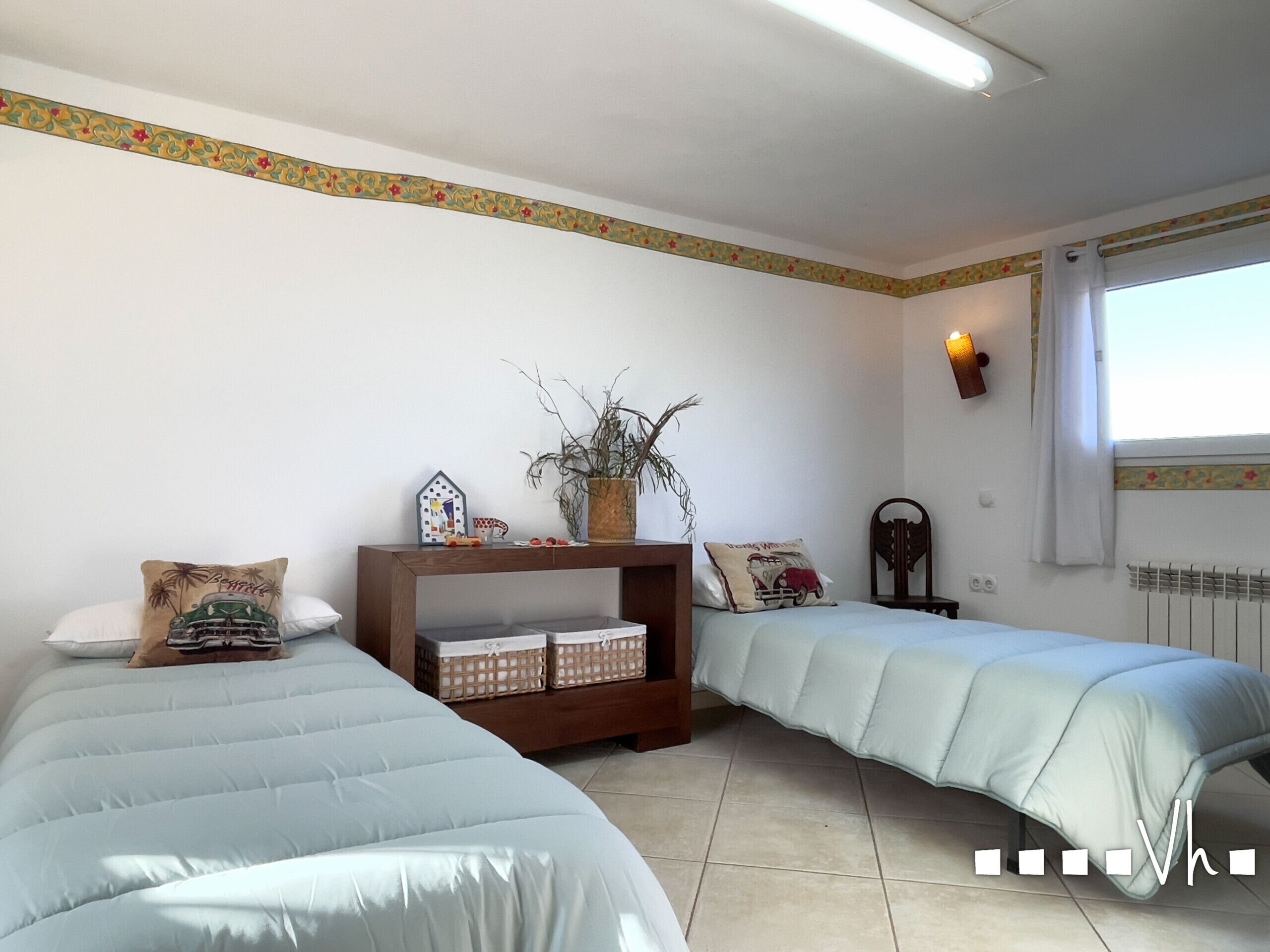 Une chambre avec deux lits, une table de chevet, une lampe fixée au mur et une fenêtre avec des rideaux.