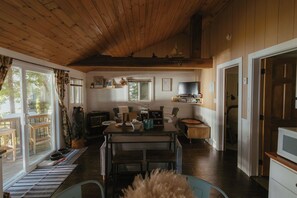 A cozy cabin interior with a wooden ceiling, a dining area with a table and chairs, a television, and a view of the outdoors through large windows.