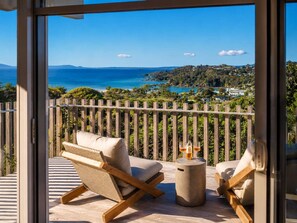 A balcony with wicker chairs, a small round table, and a view of the sea and landscape. - Oneroa Vines and Views walk to Oneroa Village (Waiheke Island)