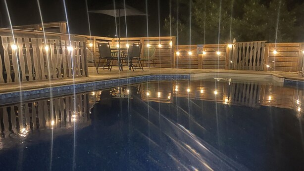 A nighttime swimming pool with a wooden deck and chairs, surrounded by a wooden fence with lights.