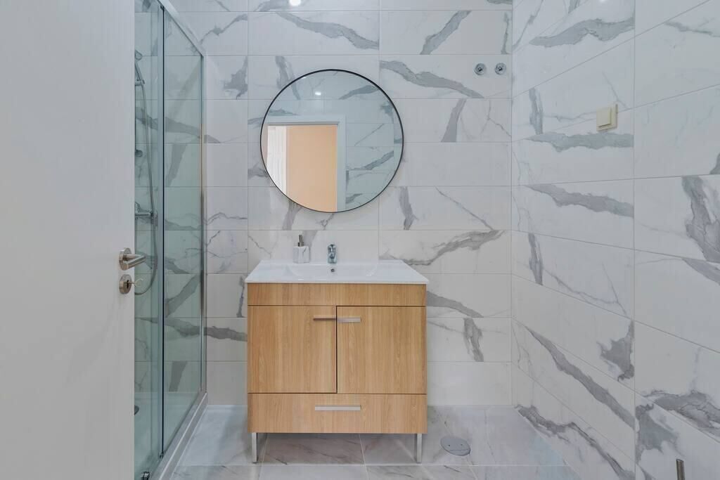 A modern bathroom with a glass shower enclosure, a wooden vanity with a round mirror, and marble-patterned walls.