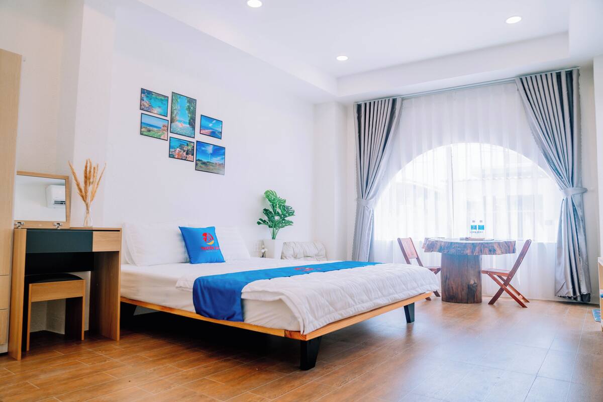 Una habitación de hotel moderna con una cama de madera, un escritorio, una silla y una mesa de comedor. Hay cuatro cuadros en la pared y una planta en maceta.