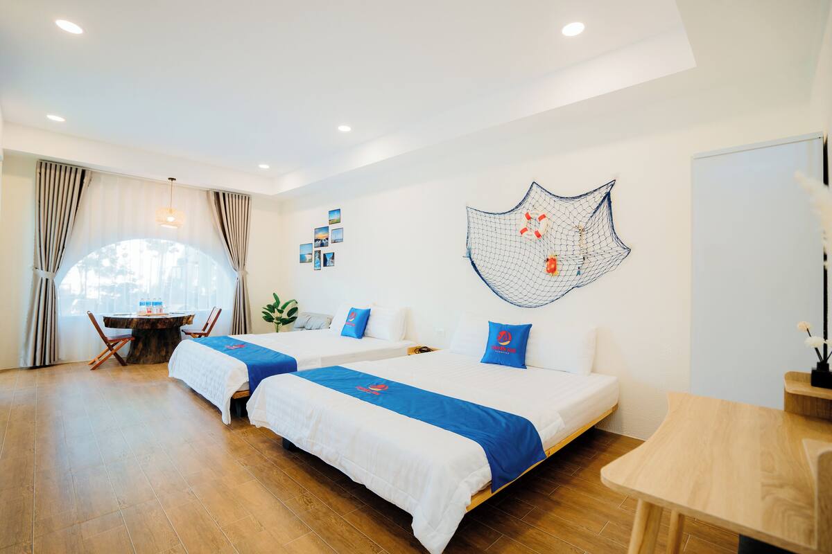 Una habitación de hotel con dos camas, una mesa redonda y un ventanal grande con cortinas.
